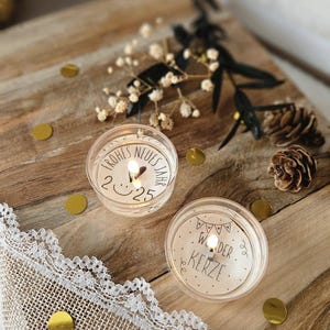 May include: Two small, clear glass votive candles with white labels. The first candle has the text "Frohes Neues Jahr 2025" on the label. The second candle has the text "Wunder Kerze" on the label. The candles are sitting on a wooden surface with gold confetti.