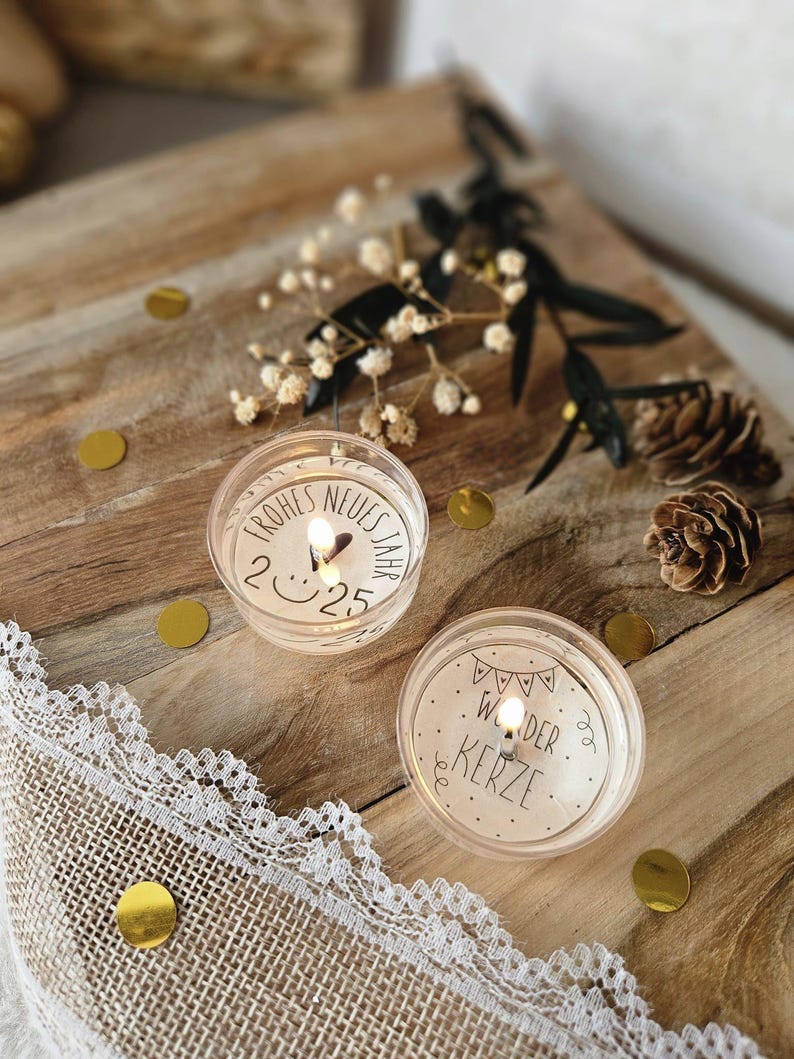 May include: Two small, clear glass votive candles with white labels. The first candle has the text "Frohes Neues Jahr 2025" on the label. The second candle has the text "Wunder Kerze" on the label. The candles are sitting on a wooden surface with gold confetti.