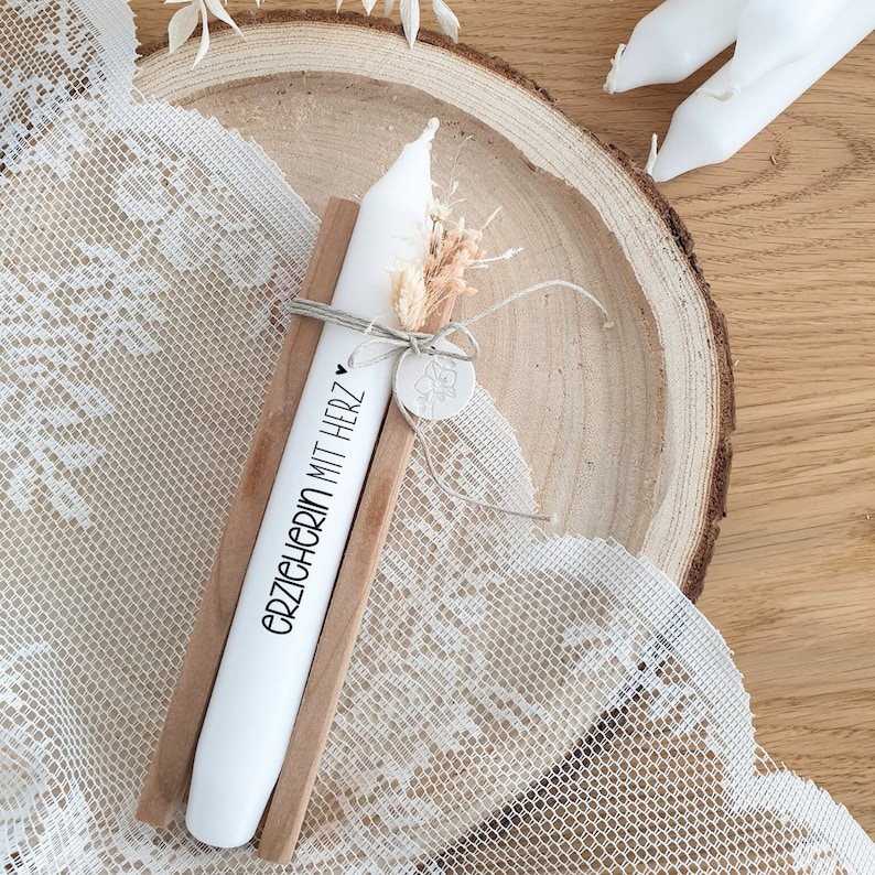 Puede incluir: Una vela blanca con el texto "erzichten mit herz" en ella, colocada en un soporte de madera. La vela est&aacute; decorada con flores secas y una peque&ntilde;a etiqueta de arcilla.