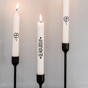 K&ouml;nnte beinhalten: Drei wei&szlig;e Kerzen mit schwarzer Beschriftung und einem Kleeblatt-Design auf schwarzen Kerzenst&auml;ndern. Die Kerzen sind angez&uuml;ndet und der Text auf einer Kerze lautet "Gl&uuml;ckwunsch".