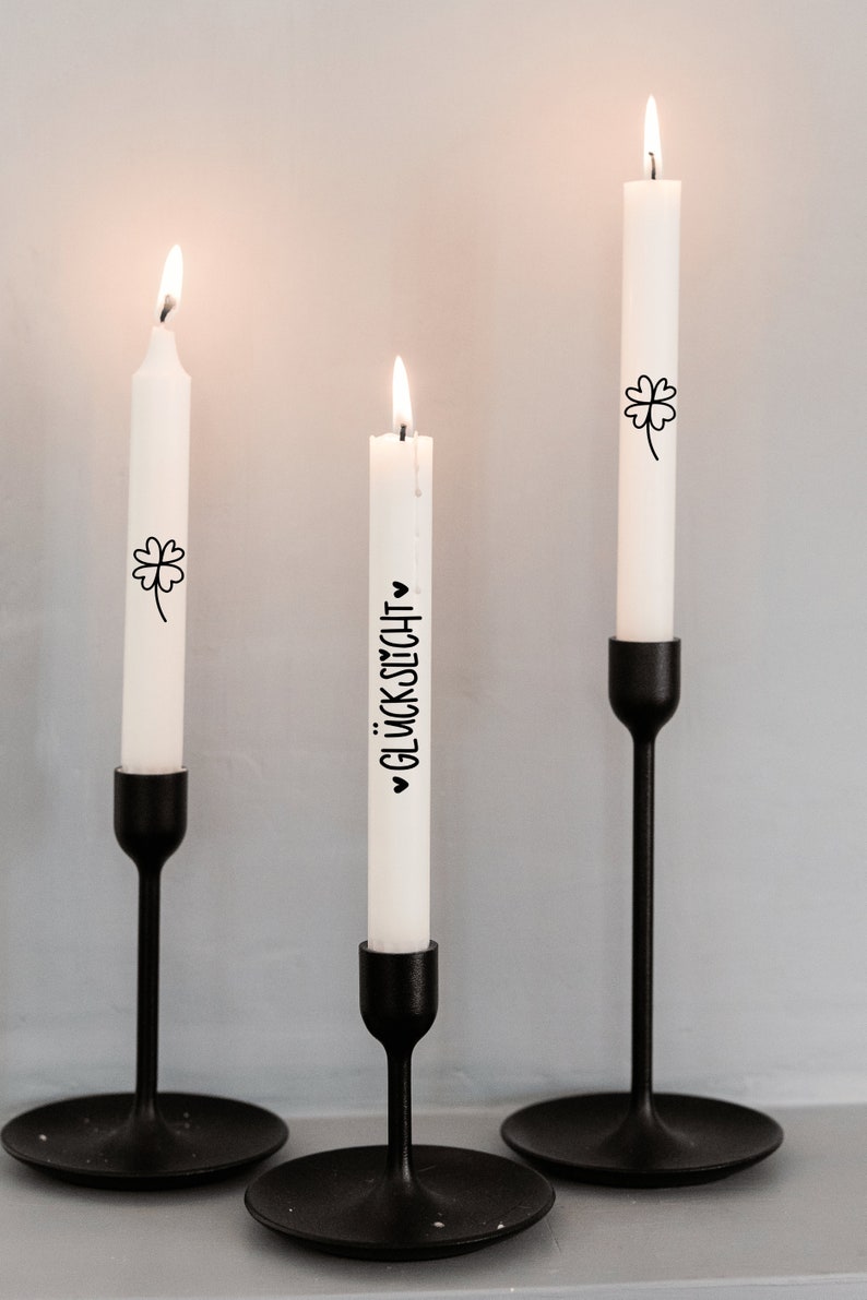 K&ouml;nnte beinhalten: Drei wei&szlig;e Kerzen mit schwarzer Beschriftung und einem Kleeblatt-Design auf schwarzen Kerzenst&auml;ndern. Die Kerzen sind angez&uuml;ndet und der Text auf einer Kerze lautet "Gl&uuml;ckwunsch".