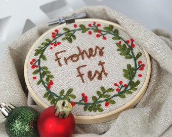 May include: A round embroidery hoop with a white linen background and a green and red embroidered wreath with the text "Frohes Fest" in brown cursive lettering.
