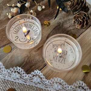 May include: Two white tea light candles with black text. The first candle says "Frohes Neues Jahr 2025" with a smiley face. The second candle says "Wunder Kerze" with a string of flags.