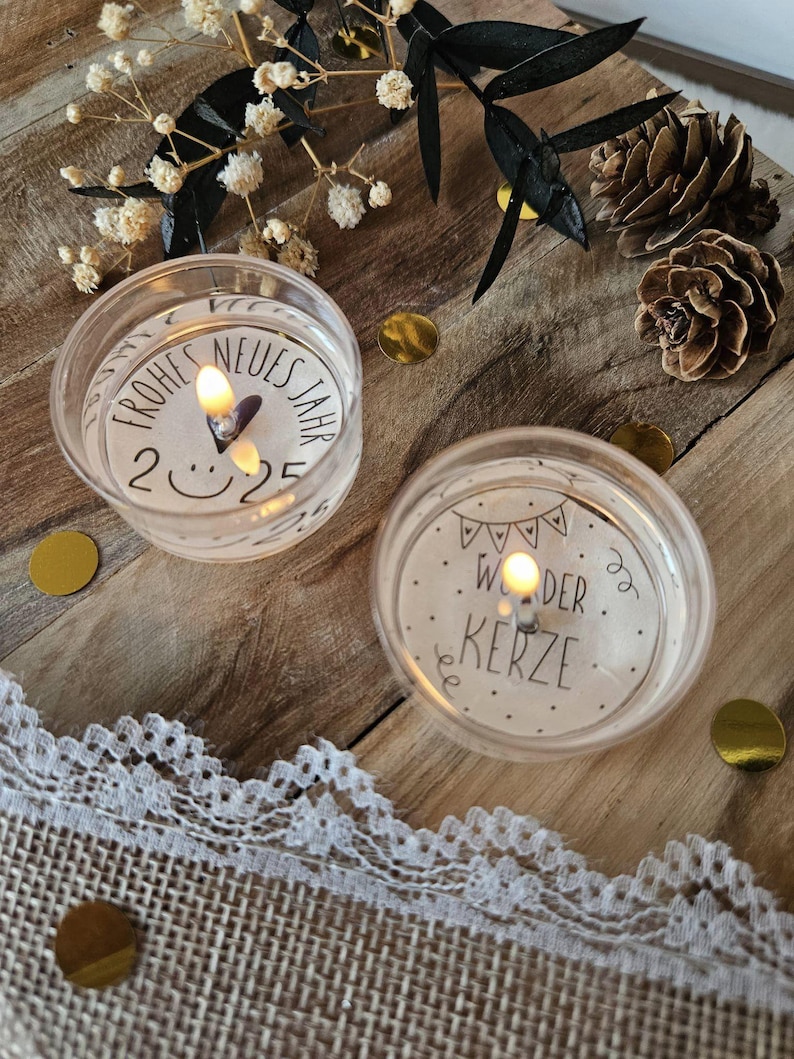 May include: Two white tea light candles with black text. The first candle says "Frohes Neues Jahr 2025" with a smiley face. The second candle says "Wunder Kerze" with a string of flags.