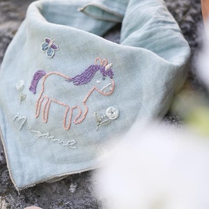 Può includere: Un bandana azzurro con dettagli ricamati. Il bandana presenta un design a unicorno con capelli viola e un corno, insieme alla parola "magical". Ulteriori elementi ricamati includono una farfalla e fiori. Il bandana è realizzato in tessuto morbido.