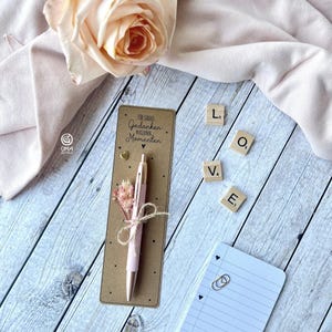 May include: A pink pen with gold detailing sits on a brown card inscribed with the German phrase "Für große Gedanken in kleinen Momenten." The card features a small gold heart and dried flowers. Wooden letter tiles spell "LOVE".