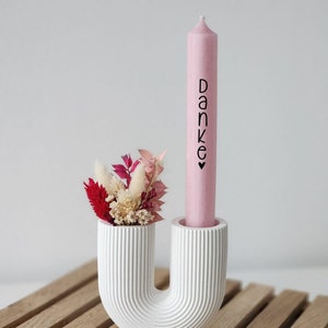 Puede incluir: Un portavelas de cer&aacute;mica blanca con forma de U con una vela rosa que dice "Danke" en letras negras. El portavelas tiene un peque&ntilde;o ramo de flores secas en la parte superior.
