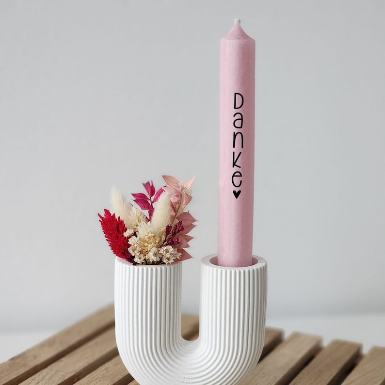 Puede incluir: Un portavelas de cer&aacute;mica blanca con forma de U con una vela rosa que dice "Danke" en letras negras. El portavelas tiene un peque&ntilde;o ramo de flores secas en la parte superior.
