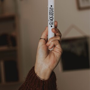 K&ouml;nnte beinhalten: Eine wei&szlig;e Kerze mit einer schwarzen Aufschrift "&hearts; GL&Uuml;CKSLICHT &hearts;" wird von einer Hand gehalten. Die Kerze ist angez&uuml;ndet und die Flamme ist sichtbar.