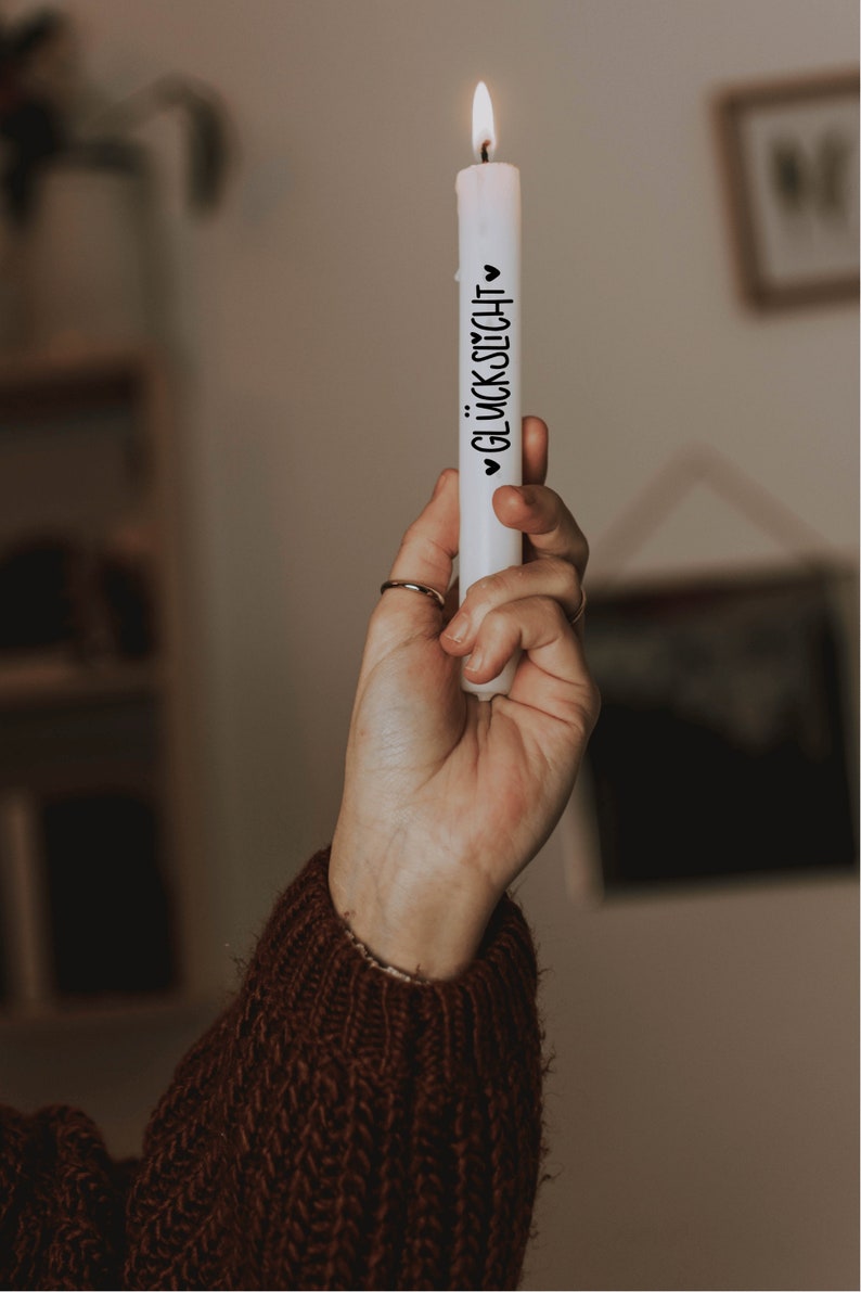 K&ouml;nnte beinhalten: Eine wei&szlig;e Kerze mit einer schwarzen Aufschrift "&hearts; GL&Uuml;CKSLICHT &hearts;" wird von einer Hand gehalten. Die Kerze ist angez&uuml;ndet und die Flamme ist sichtbar.