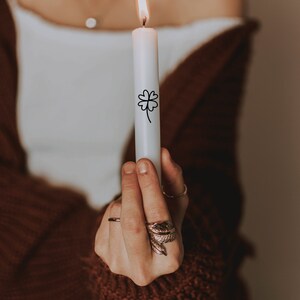 K&ouml;nnte beinhalten: Eine Person h&auml;lt eine wei&szlig;e Kerze mit einem schwarzen vierbl&auml;ttrigen Kleeblatt-Design. Die Kerze ist angez&uuml;ndet. Die Person tr&auml;gt einen braunen Pullover.