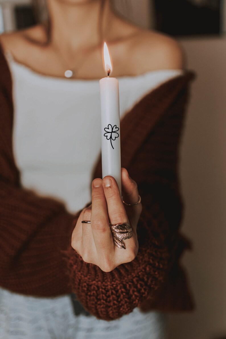 K&ouml;nnte beinhalten: Eine Person h&auml;lt eine wei&szlig;e Kerze mit einem schwarzen vierbl&auml;ttrigen Kleeblatt-Design. Die Kerze ist angez&uuml;ndet. Die Person tr&auml;gt einen braunen Pullover.