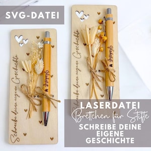 May include: Two wooden plaques with a pen and dried flowers. The plaques are engraved with the text "Schreibe deine eigene Geschichte" which translates to "Write your own story". The pen is engraved with the text "Glocksstift" which translates to "Good luck pen".