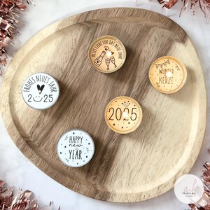 May include: Four round tea light candles with different New Year's Eve designs. The candles are on a wooden tray. The designs include "Happy New Year", "Frohes Neues Jahr 2025", "Alles Liebe fur das Neue Jahr", and "Wunder Kerze".