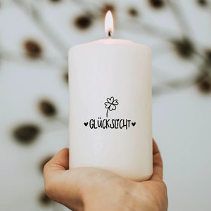 K&ouml;nnte beinhalten: Eine wei&szlig;e Kerze mit einem vierbl&auml;ttrigen Kleeblatt und dem Text "Gl&uuml;cklicht" in schwarzer Schrift. Die Kerze ist angez&uuml;ndet und wird von einer Hand gehalten.