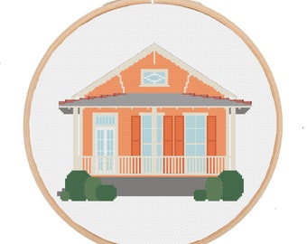 May include: A cross-stitch pattern of a peach-colored house with a gray roof, white trim, and blue windows. The house has a porch with white railings and green bushes on either side.