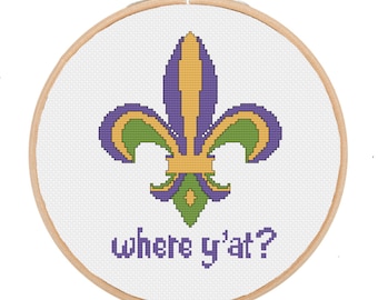 May include: A cross-stitch embroidery in a wooden hoop features a fleur-de-lis design in purple, green, and gold. The text "where y'at?" is stitched below the emblem. The embroidery is on a white background.