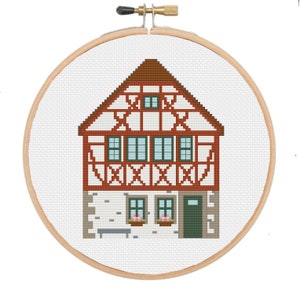 May include: Cross-stitch embroidery of a traditional European house, framed in a wooden hoop. The house features a brown roof, red and white timber framing, green window frames, and a gray stone base. The design is on a white fabric background.
