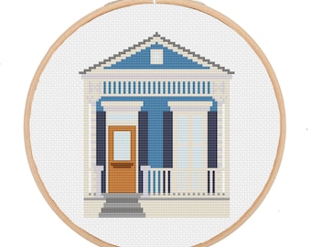 May include: Cross-stitch embroidery of a house in a wooden hoop. The house features a blue roof, white trim, and a brown door. The design is set against a white background, creating a charming, handcrafted aesthetic. The hoop is approximately 6 inches in diameter.