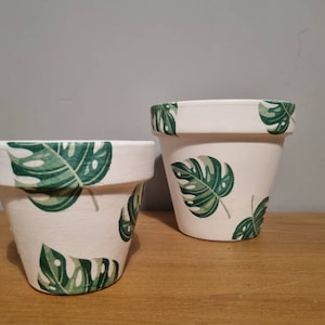 May include: Two white ceramic plant pots with a green tropical leaf pattern. The larger pot is taller than the smaller pot.