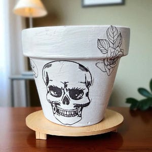 May include: White ceramic plant pot with a hand-drawn skull design. The pot has a textured surface and is decorated with black floral accents. It sits on a round, wooden plant stand. A framed skull drawing is visible in the background.