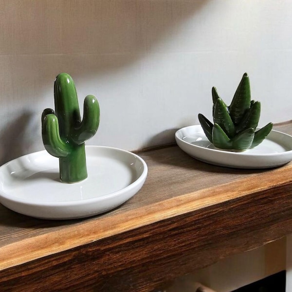 May include: Two white ceramic dishes with green ceramic cactus and succulent plants. The dishes are on a dark brown wooden surface.