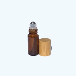 Puede incluir: Un pequeño frasco roll-on de vidrio ámbar con una bola rodante plateada y una tapa cilíndrica de bambú separada. El frasco está diseñado para aceites esenciales o perfumes. La tapa es de bambú de color claro.