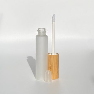 May include: A frosted glass bottle with a bamboo lid and a white applicator wand. The bottle is empty and ready to be filled with lip gloss or other liquid cosmetics.