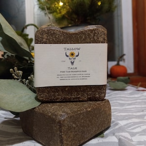 May include: Two dark brown, rectangular shampoo bars are stacked. The top bar has a white label with the text "Tallow Soap Co." and "TALK Pine Tar Shampoo Bar." The bars are on a patterned surface with greenery in the background.