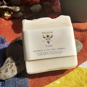 May include: A bar of soap with a paper label wrapped around it. The label features a sunflower and bull skull logo and the words "TALLOW SOAP CO". The text "TALK ROSEMARY & TEA TREE SHAMPOO" is also visible. The soap is on a patterned surface.