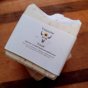 Könnte beinhalten: Ein rechteckiges Seifenstück, verpackt in einem weißen Etikett. Das Etikett trägt den Text "TALLOW TALK CO." und "NETTLE + CLARY SAGE SHAMPOO BAR." Die Seife hat eine helle Farbe mit sichtbaren Flocken. Der Hintergrund ist eine Holzoberfläche.