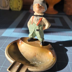 May include: Vintage ashtray featuring a small figurine of a man in overalls and a hat. The ashtray is a gold color with a small rest for cigarettes. The figurine has a white shirt, green overalls, and a gray hat.