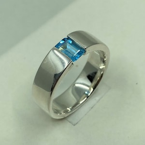 Könnte beinhalten: Ein polierter Silberring mit einem rechteckigen, hellblauen Edelstein. Das Band ist breit und glatt und reflektiert das Licht. Der Ring wird auf einer weißen Oberfläche präsentiert, um sein Design und seine Farbe hervorzuheben.