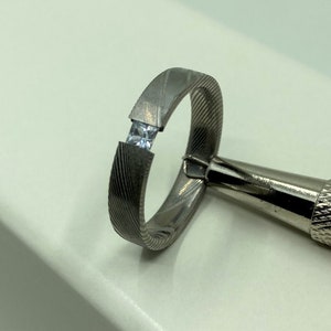 Könnte beinhalten: Ein silberner Ring mit einem einzigen, klaren, quadratischen Edelstein, der in die Band gefasst ist. Der Ring hat eine strukturierte, gebürstete Oberfläche.