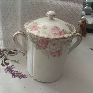 May include: A white porcelain sugar bowl with a lid, featuring a floral design with pink roses and green leaves. The bowl has two handles and a gold trim around the top.