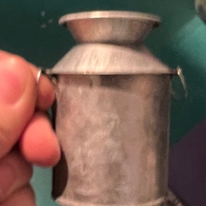 May include: A small, vintage-style metal milk can with a lid and handle. The can is silver-colored and appears to be made of tin or a similar metal. The design is simple and functional, suggesting it may be a decorative item or a miniature collectible.