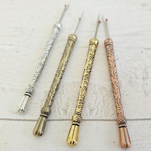 May include: Four decorative seam rippers in silver, brass, gold, and copper. Each ripper features an ornate, engraved handle and a pointed blade with a red tip. The handles have a textured, patterned design.