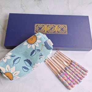 May include: A light blue zipped pouch featuring a sunflower and bee pattern, accompanied by a set of eight crochet hooks with floral patterned handles, all presented in a dark blue rectangular box with the gold text Gift Set on the lid.