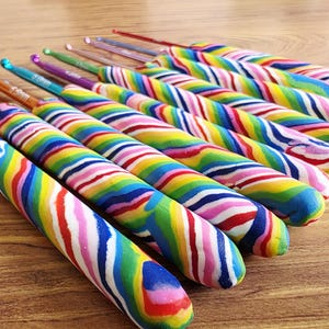 May include: A set of crochet hooks featuring metallic tips in various colours and thick, ergonomic handles with a swirling, multi-coloured rainbow pattern. Each hook displays its size clearly on the metal shaft, arranged in a row on a surface.