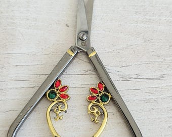 May include: Decorative silver scissors with gold accents and jewel-like embellishments. The handles feature a floral design with red and green gemstones. The scissors are open, displaying the sharp blades.