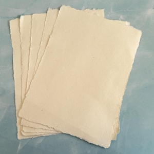 May include: A stack of rectangular sheets of off-white paper with uneven, deckled edges. The paper has a visible fibrous texture and a matte finish, layered slightly offset to reveal multiple individual sheets in the collection.