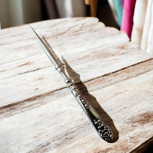 May include: An antique letter opener with a silver-colored blade and ornate handle. The handle features detailed patterns and a decorative end. The opener is resting on a weathered, light-colored wooden surface.
