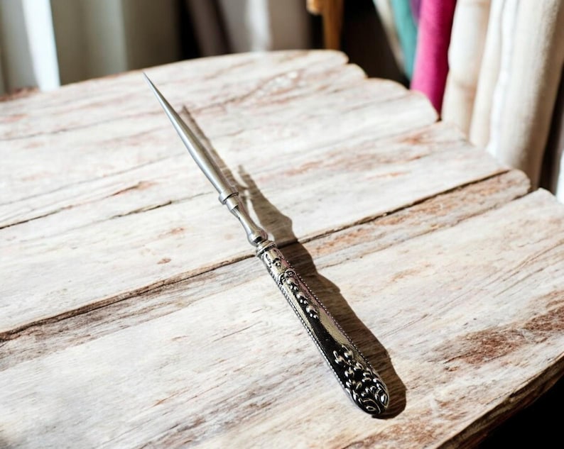 May include: An antique letter opener with a silver-colored blade and ornate handle. The handle features detailed patterns and a decorative end. The opener is resting on a weathered, light-colored wooden surface.