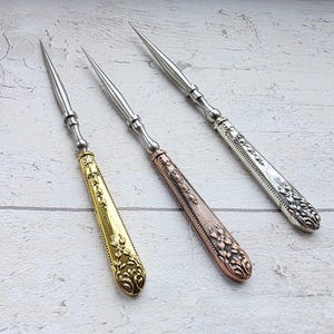 May include: Three ornate metal awls with pointed tips. One has a gold handle, another a copper handle, and the third a silver handle. The handles feature detailed floral and scrollwork designs, and the awls are arranged on a light wood surface.