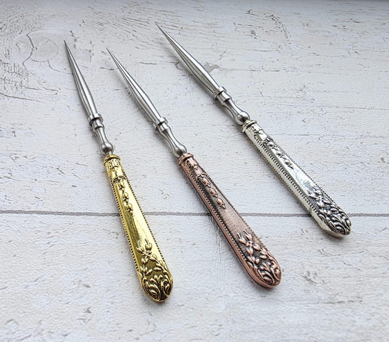 May include: Three ornate metal awls with pointed tips. One has a gold handle, another a copper handle, and the third a silver handle. The handles feature detailed floral and scrollwork designs, and the awls are arranged on a light wood surface.
