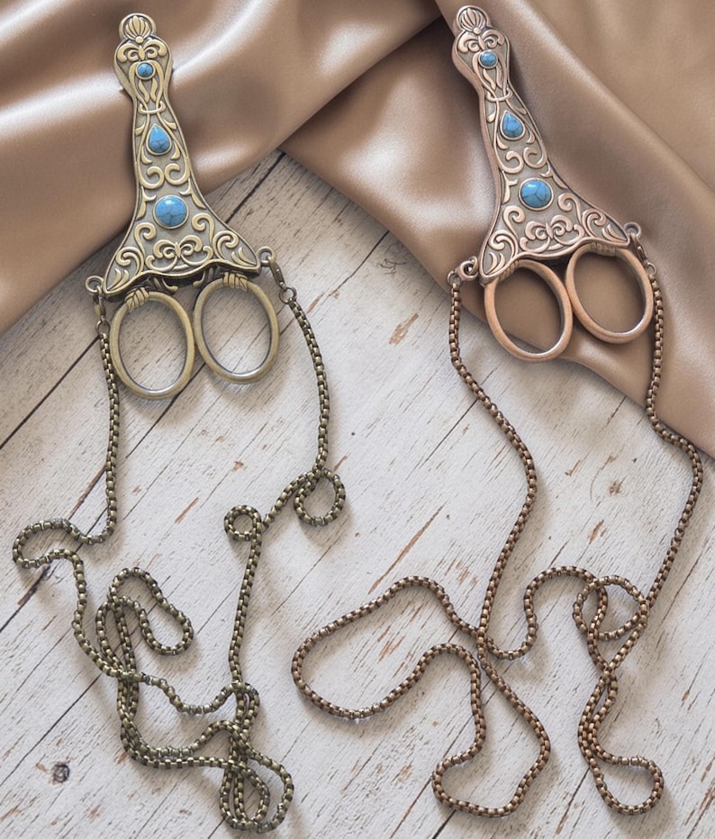 May include: Two antique-style scissors with detailed metalwork and turquoise-coloured accents. Each scissor features a decorative handle and a chain. The scissors are displayed on a light wood surface.