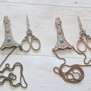 May include: Two pairs of decorative sewing scissors with ornate handles and small blades. The handles are embellished with turquoise-coloured stones and connected to a chain. One pair has a bronze finish, the other a brass finish.