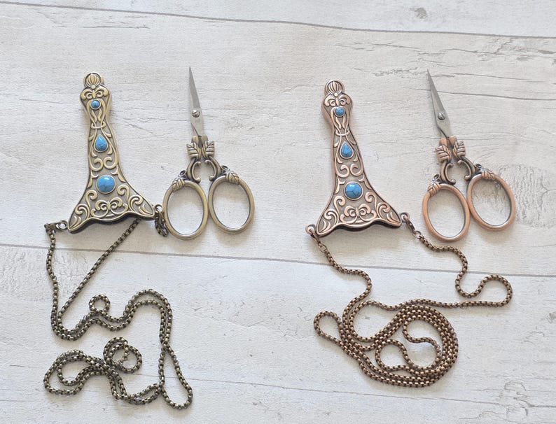 May include: Two pairs of decorative sewing scissors with ornate handles and small blades. The handles are embellished with turquoise-coloured stones and connected to a chain. One pair has a bronze finish, the other a brass finish.