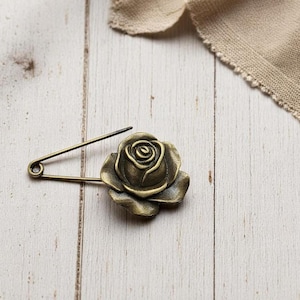 May include: A bronze-coloured safety pin featuring a rose-shaped decoration. The pin is set against a light-coloured, weathered wooden surface, with a piece of beige fabric in the background. The rose has detailed petals.