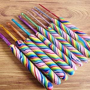 May include: A set of nine crochet hooks with colourful, swirled, multi-coloured handles. Each metal hook features a specific size printed on the shaft, ranging from 2.5mm to 6.5mm. The hooks are arranged in a diagonal row, showcasing various metallic colours.